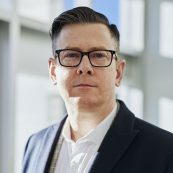 a man in a suit and glasses standing in front of a window.